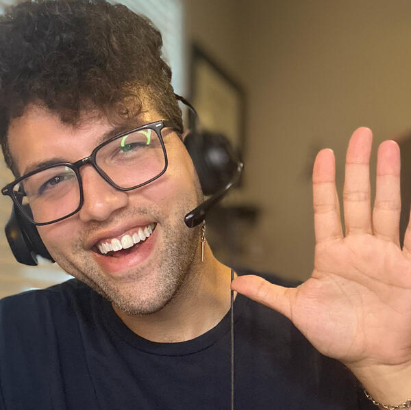 Friendly ESL teacher wearing glasses and a headset smiles and waves at the camera during an online lesson, seated indoors in a softly lit room.